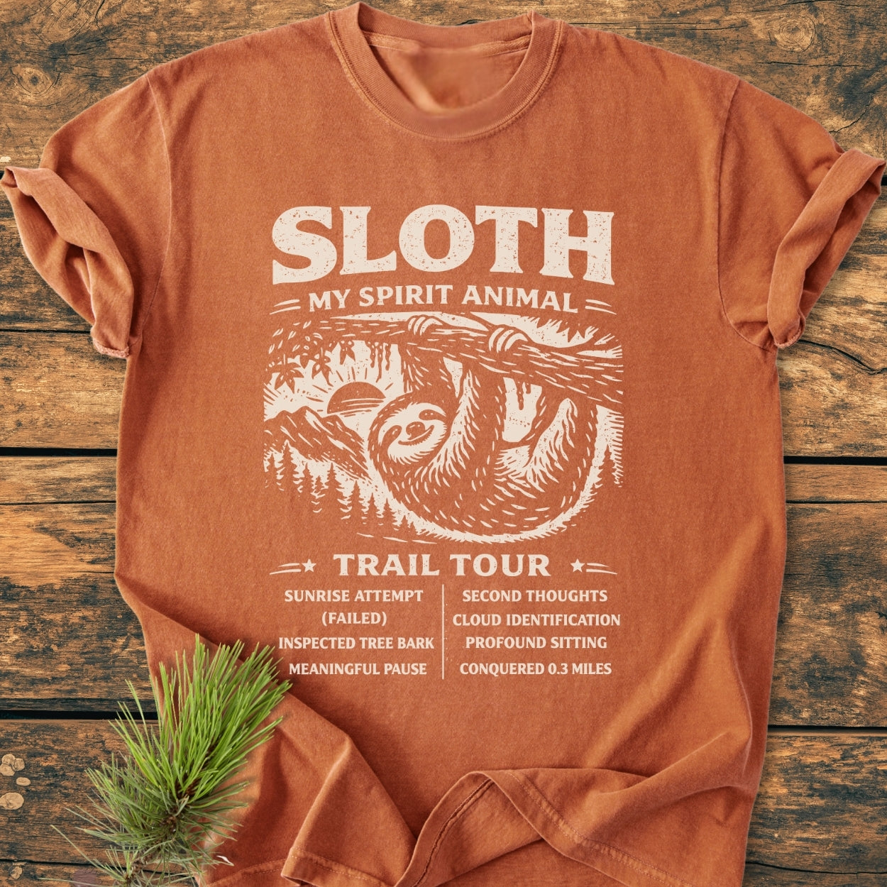 Sloth Spirit Animal Tour - Vintage Wash Tee, featuring a smiling sloth illustration and quirky hiking highlights, laid flat on a wooden surface. Made of soft, durable, pre-shrunk 100% ring-spun cotton.
