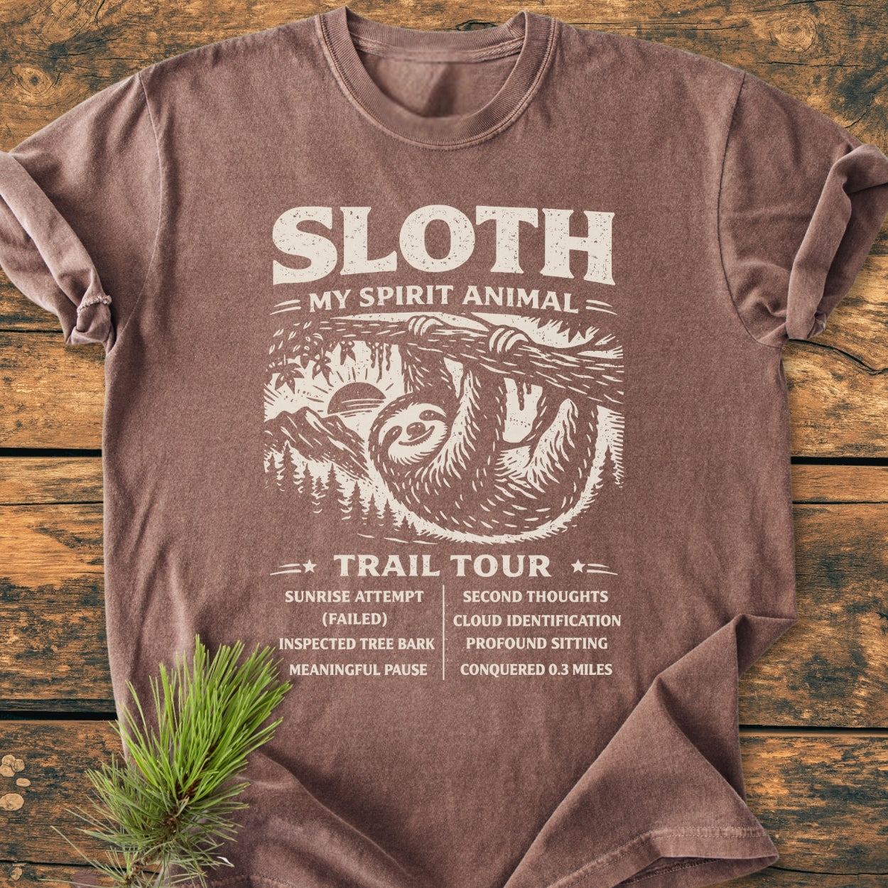 Sloth Spirit Animal Tour - Vintage Wash Tee featuring a humorous sloth design with trail accomplishments, laid flat on a wooden surface, showcasing its relaxed fit and worn-look appeal.