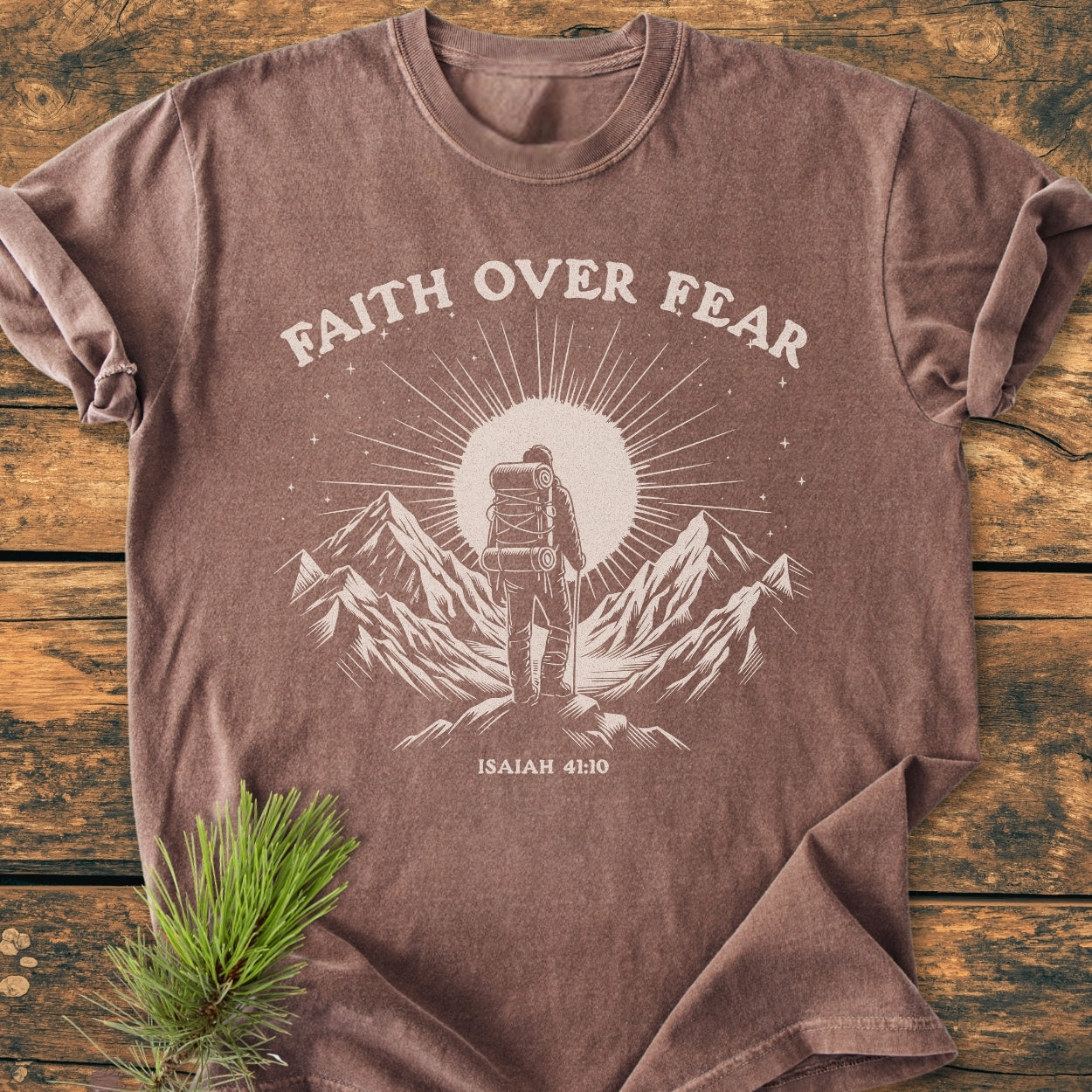 Faith Over Fear - Vintage Wash Tee featuring a brown short-sleeved design with Faith Over Fear slogan, a backpacker illustration, and Isaiah 41:10 text, displayed on a wooden surface with pine sprig.