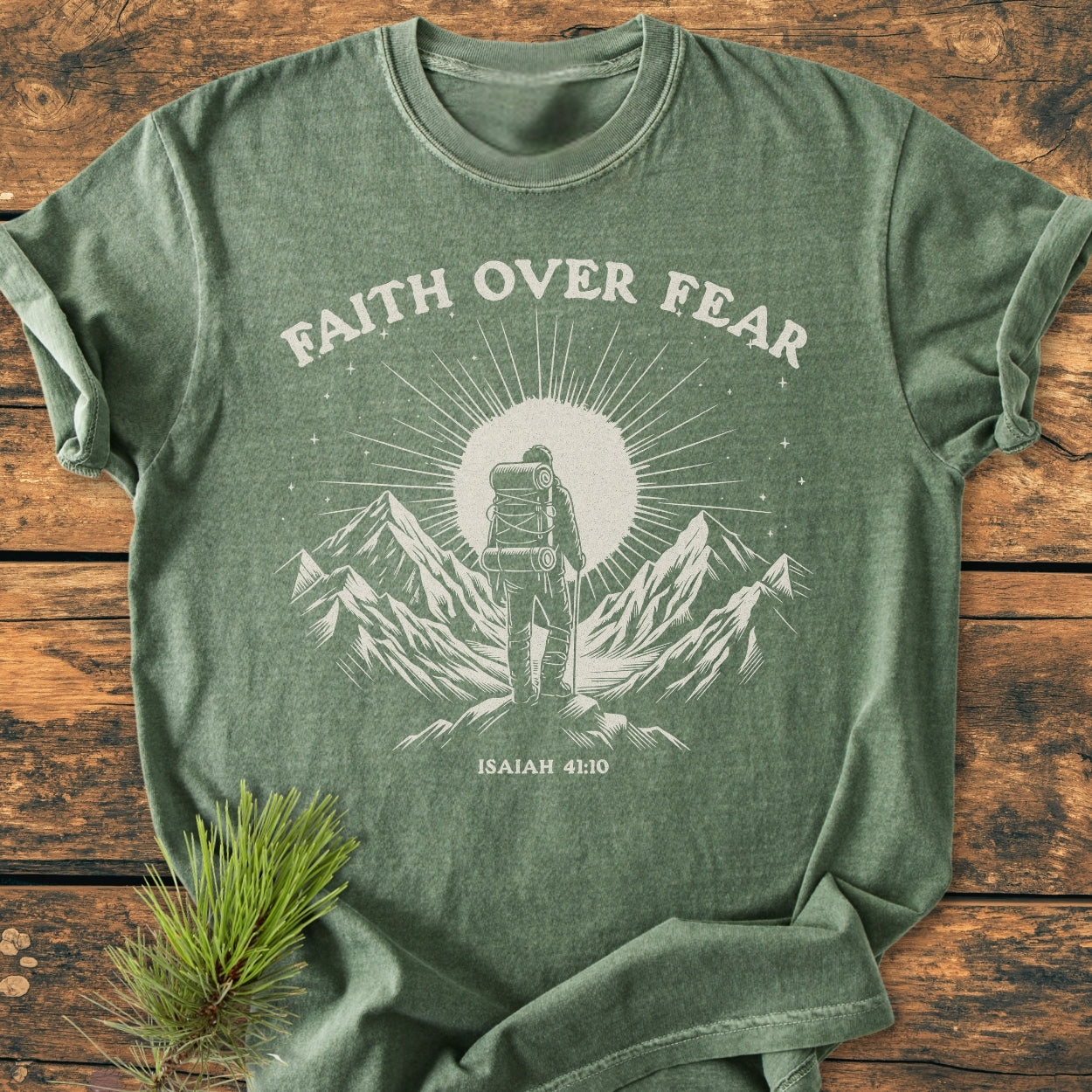 Faith Over Fear - Vintage Wash Tee features a white graphic of a backpacker on a peak, with Faith Over Fear text and Isaiah 41:10, on forest-green fabric, laid on rustic wood.