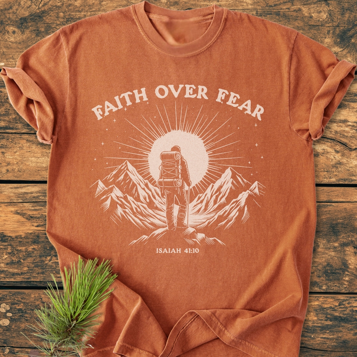 Faith Over Fear - Vintage Wash Tee featuring a white graphic of a backpacker on a mountain ridge, with bold lettering and Bible reference, displayed flat on a wooden surface with pine sprig accent.