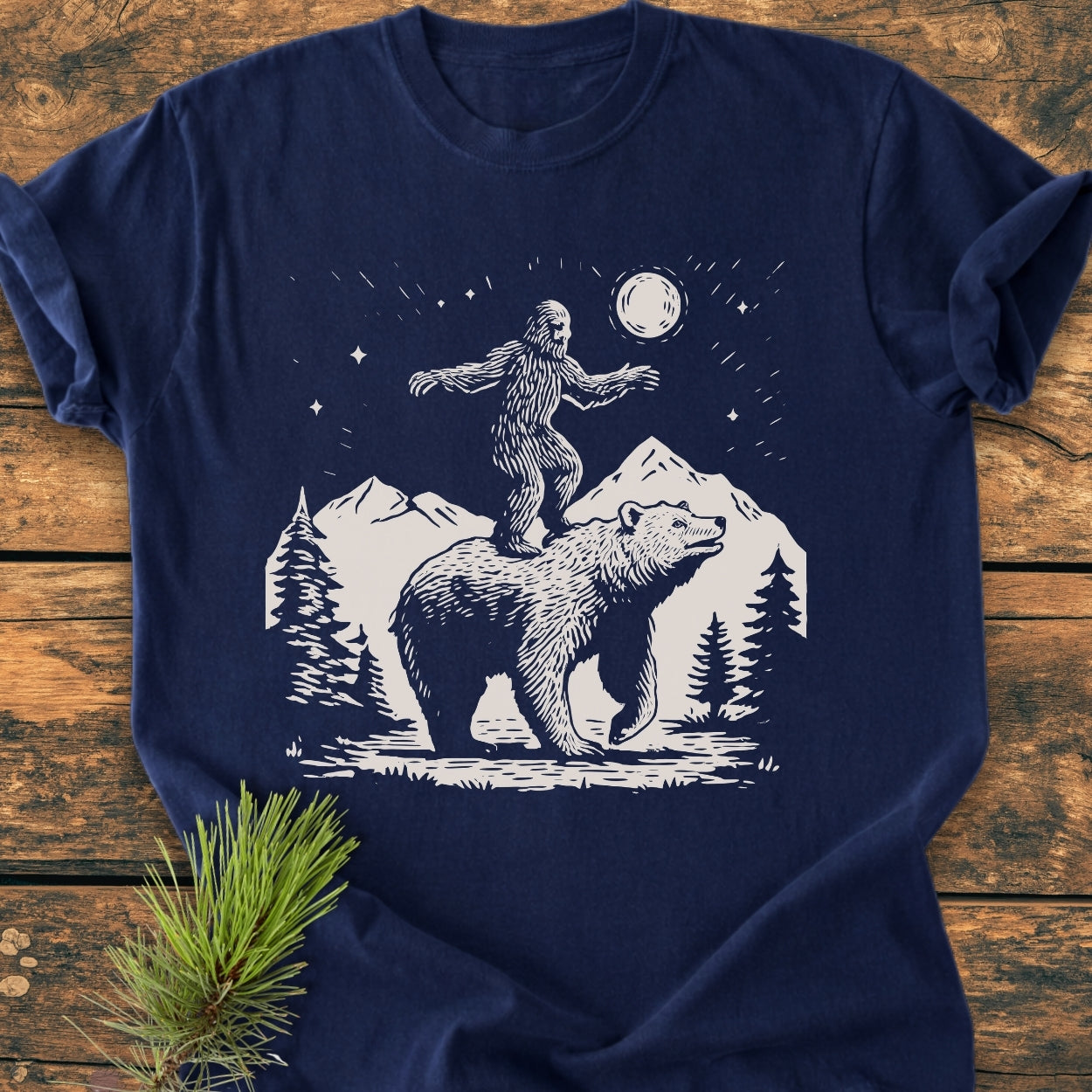 Bigfoot And Bear - Vintage Wash Tee features a navy-blue design with a stylized wilderness scene, including Bigfoot on a bear, pine trees, and a moonlit sky, enhancing its cozy, relaxed fit.