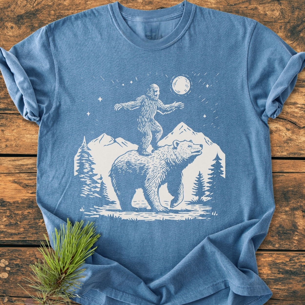 Bigfoot And Bear - Vintage Wash Tee features a woodcut-style Bigfoot and bear scene under a starry sky, laid flat on a wooden surface with a pine sprig accenting its charm.