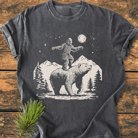 Bigfoot And Bear - Vintage Wash Tee features a stylized wilderness scene with Bigfoot and a bear, printed on a charcoal-gray T-shirt, laid on a wooden surface with a pine sprig beside it.