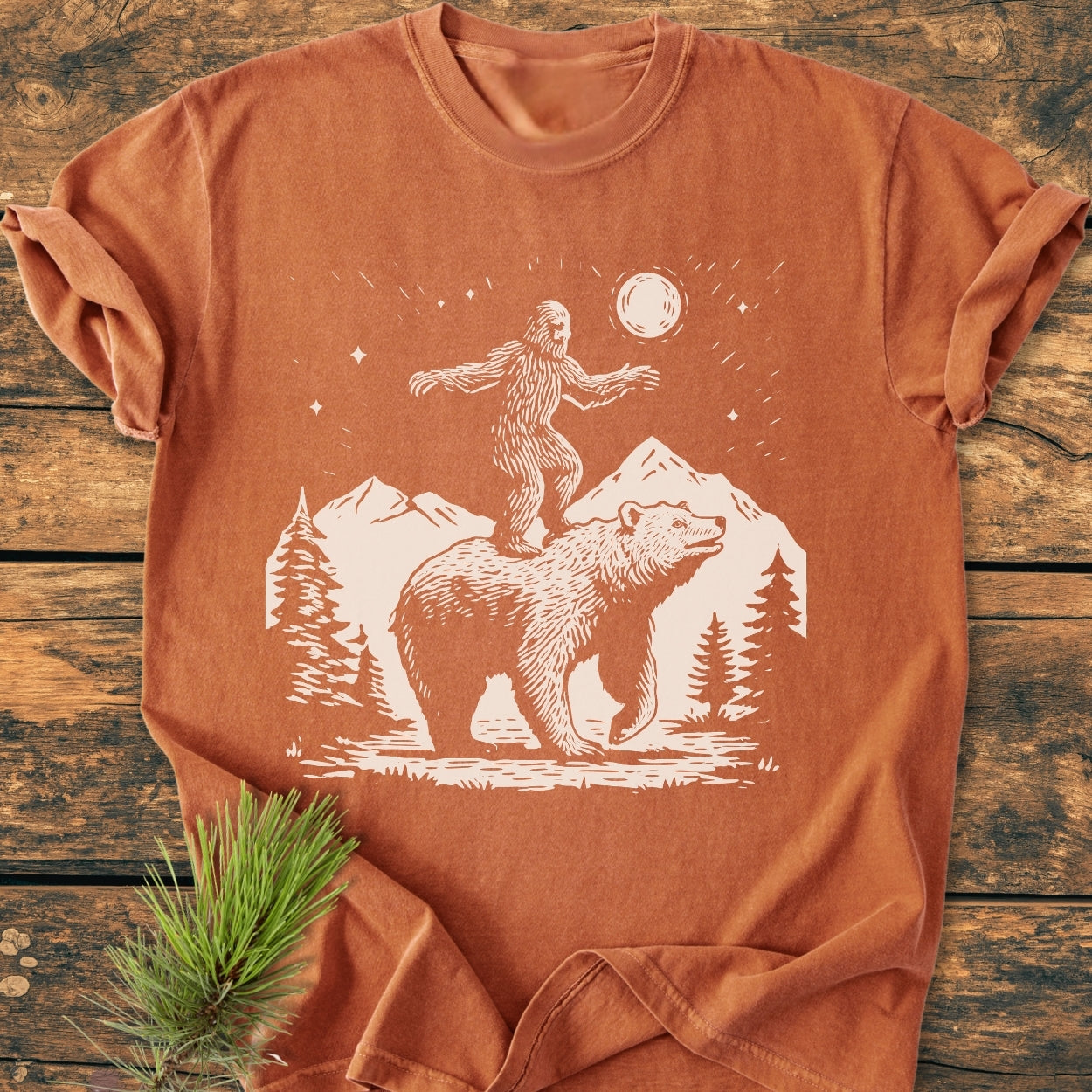 Bigfoot And Bear - Vintage Wash Tee features a wilderness scene with a bear and Sasquatch under a moon, printed on a rust-orange, relaxed fit, garment-dyed, 100% ring-spun cotton T-shirt.