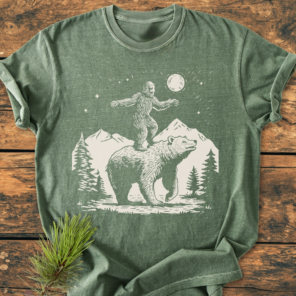 Bigfoot And Bear - Vintage Wash Tee features a woodcut-style Bigfoot scene with a bear, pine trees, mountains, and a moon on a heather-green, relaxed fit, garment-dyed crew-neck T-shirt.