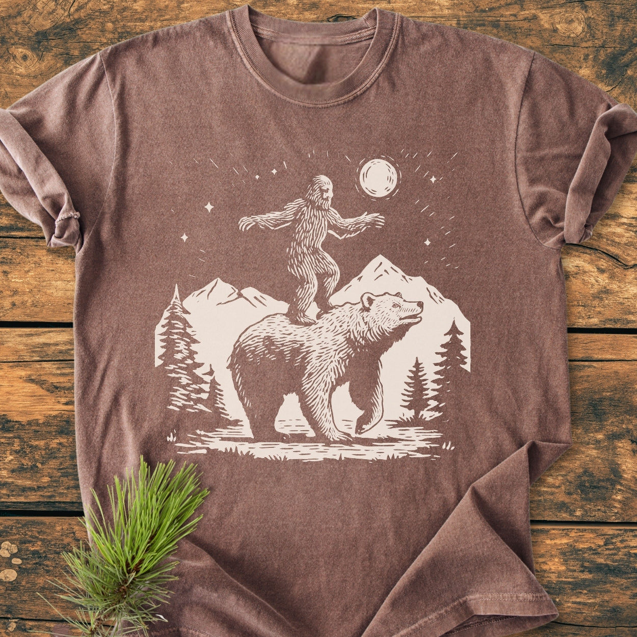 Bigfoot And Bear - Vintage Wash Tee features a stylized wilderness print with a bear, mountains, and Sasquatch. Laid flat, this relaxed, soft-washed brown tee offers comfort and durability.