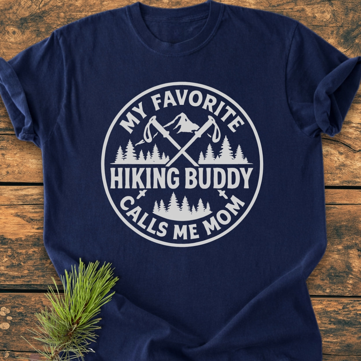 Navy-blue Hiking Buddy Mom - Vintage Wash Tee on wood surface, featuring white circular graphic with hiking poles, mountains, and trees, celebrating a mother’s hiking bond. Made from soft, durable, 100% ring-spun cotton.