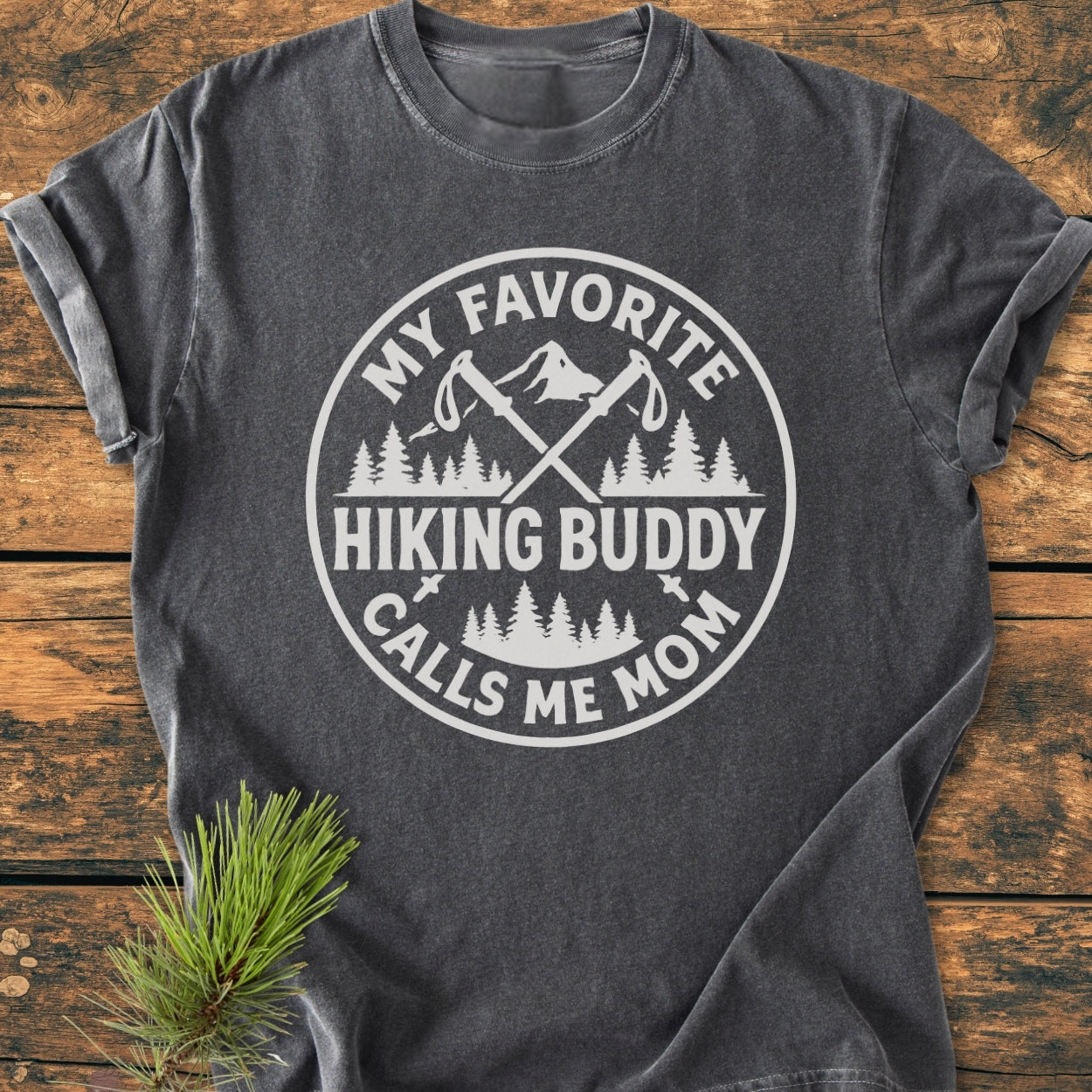 Hiking Buddy Mom - Vintage Wash Tee featuring a white circular graphic with trekking poles, pine trees, and mountain peak on a slate-gray shirt, laid on a wooden surface with pine sprigs.