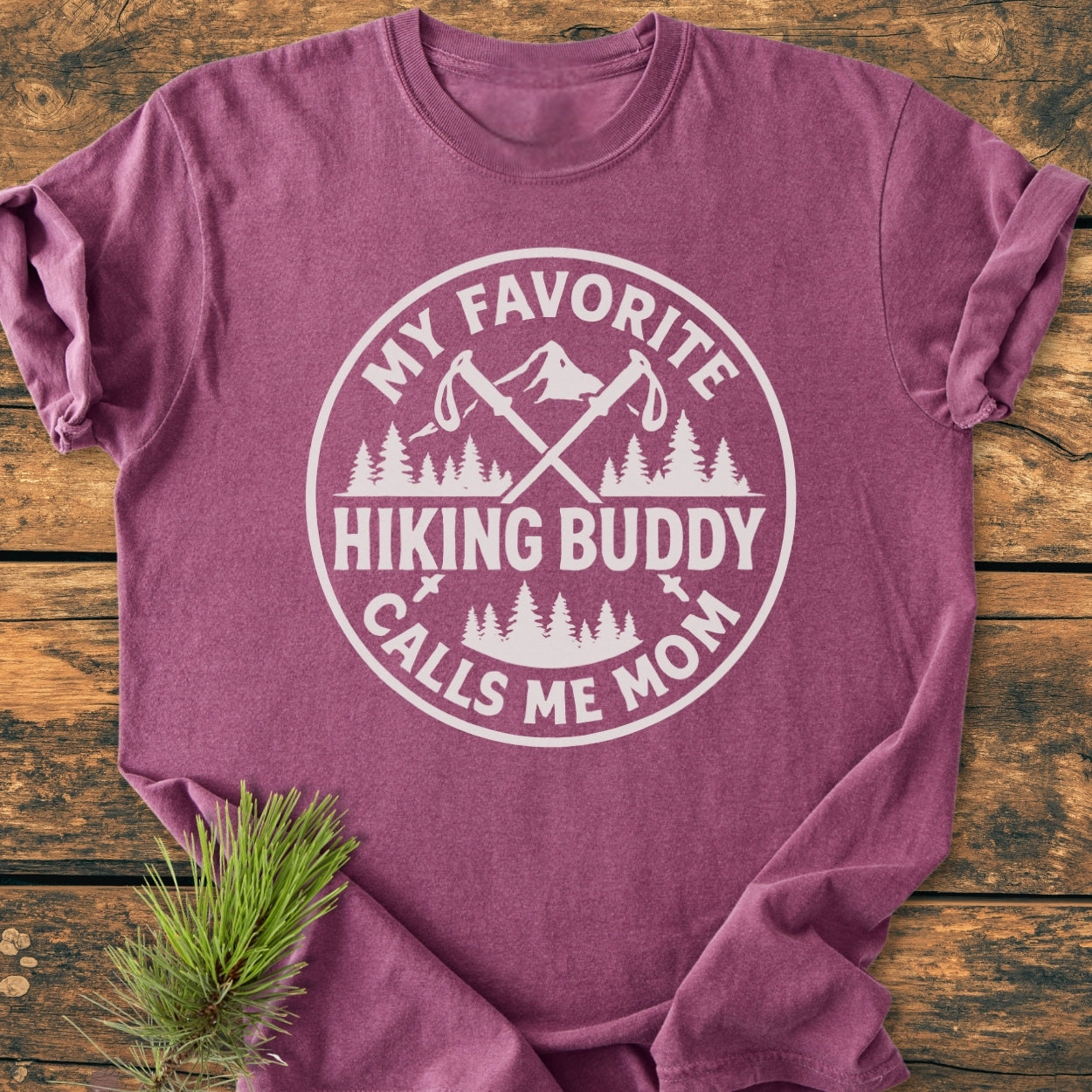 Hiking Buddy Mom - Vintage Wash Tee features a white circular graphic with crossed hiking poles, mountain, and tree line on a maroon short-sleeve T-shirt, laid on a wooden surface with a pine sprig.
