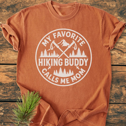 Hiking Buddy Mom - Vintage Wash Tee featuring an outdoorsy badge design with trekking poles and mountains, laid on wood-grain surface with pine sprig; a cozy, durable, relaxed-fit t-shirt.