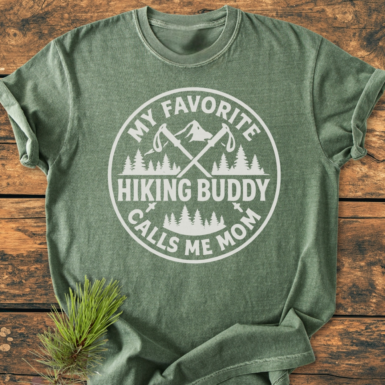 Hiking Buddy Mom - Vintage Wash Tee featuring a mountain and trees emblem with hiking poles, laid flat. Forest-green, soft-washed cotton, showcasing a relaxed fit and durable stitching, styled with a pine sprig.