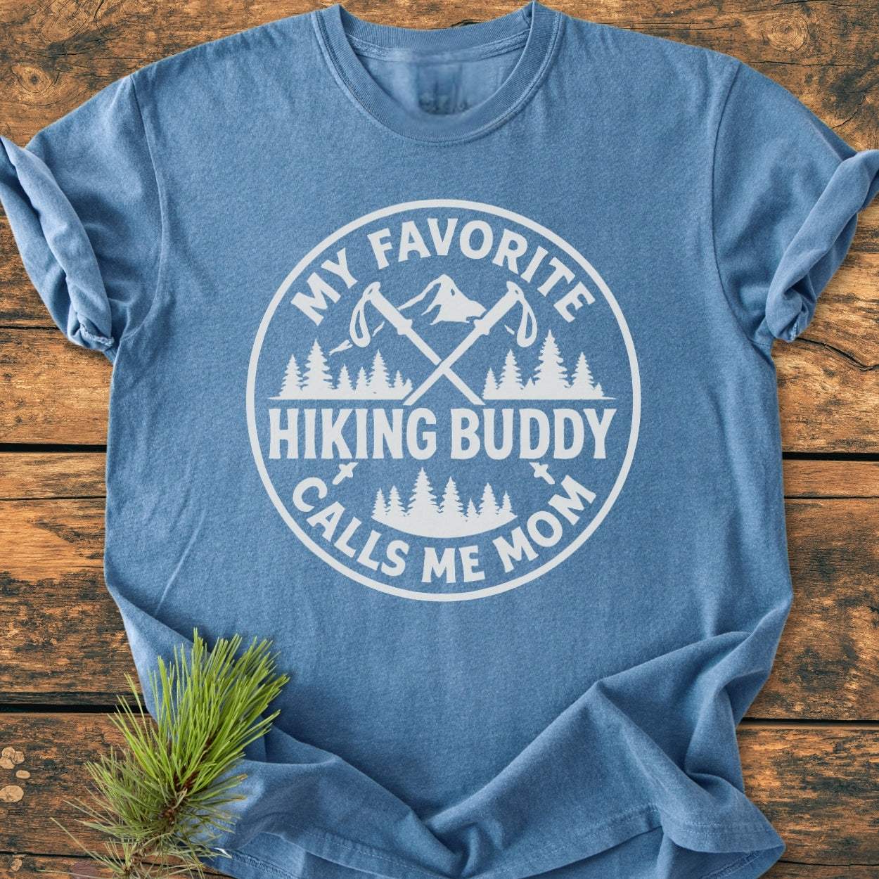 Hiking Buddy Mom - Vintage Wash Tee in heather-blue, featuring a white outdoors-themed graphic with crossed hiking poles, pine trees, and mountain peak. Laid on a wooden surface with a pine branch accent.