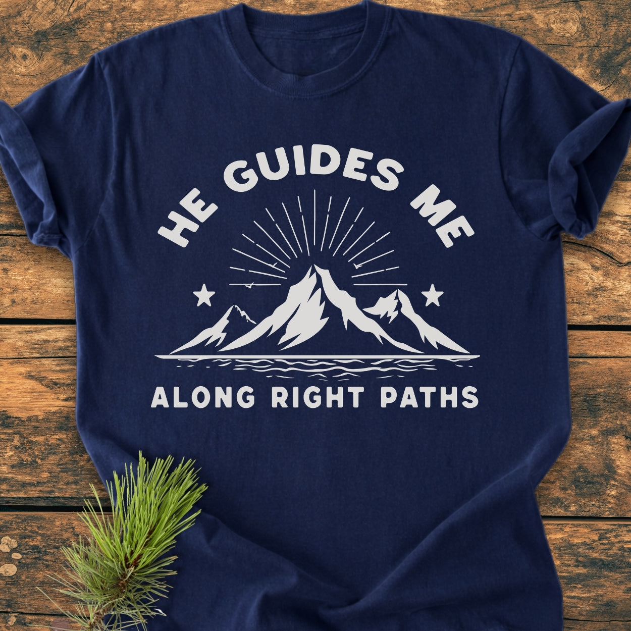He Guides Me - Vintage Wash Tee features a white stylized mountain graphic with sun rays on a navy-blue background, laid on wood. The T-shirt offers a relaxed fit with durable stitching.