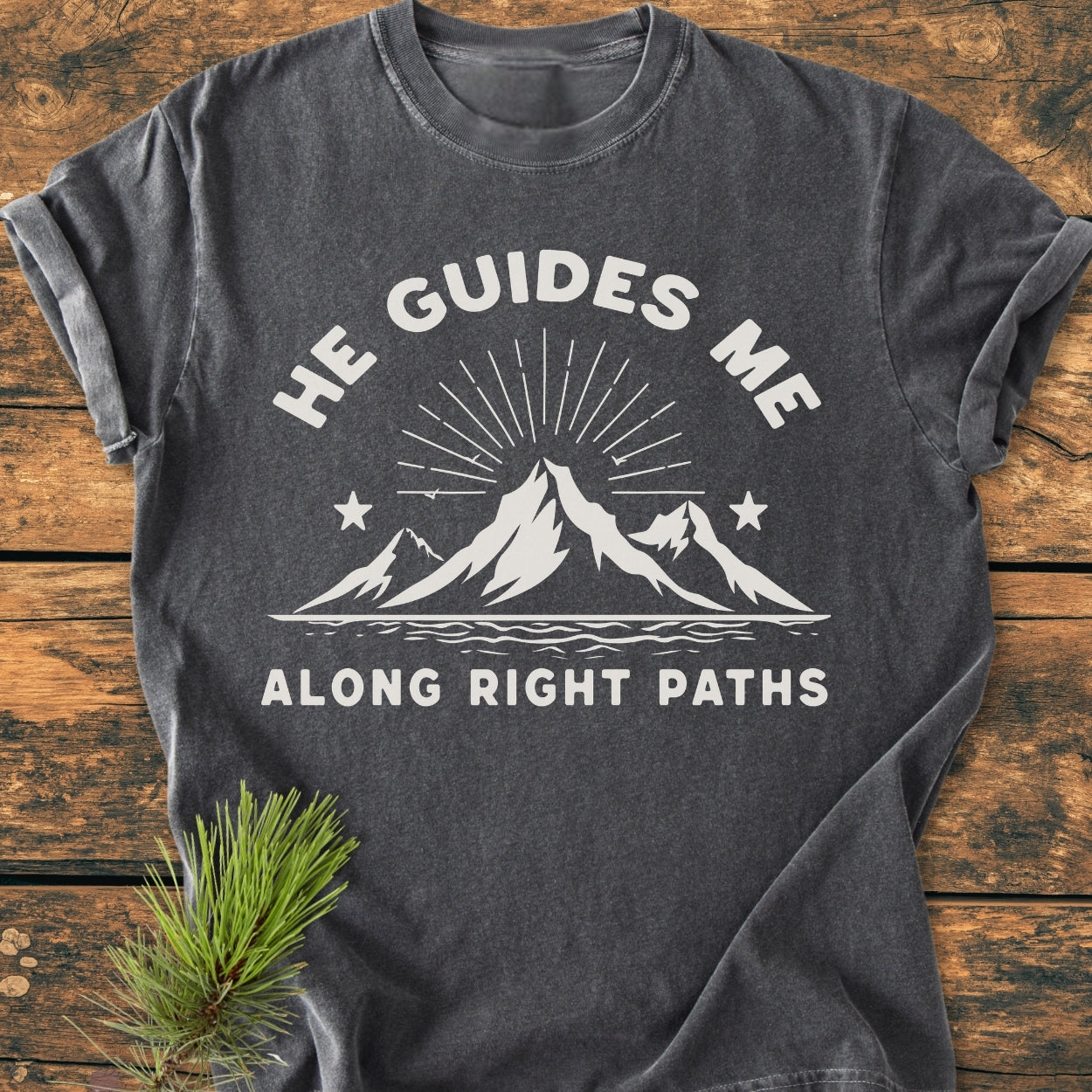 He Guides Me - Vintage Wash Tee features a charcoal-gray color with a white printed mountain design and inspirational text, laid on a wooden surface with a pine sprig, highlighting its rustic charm.