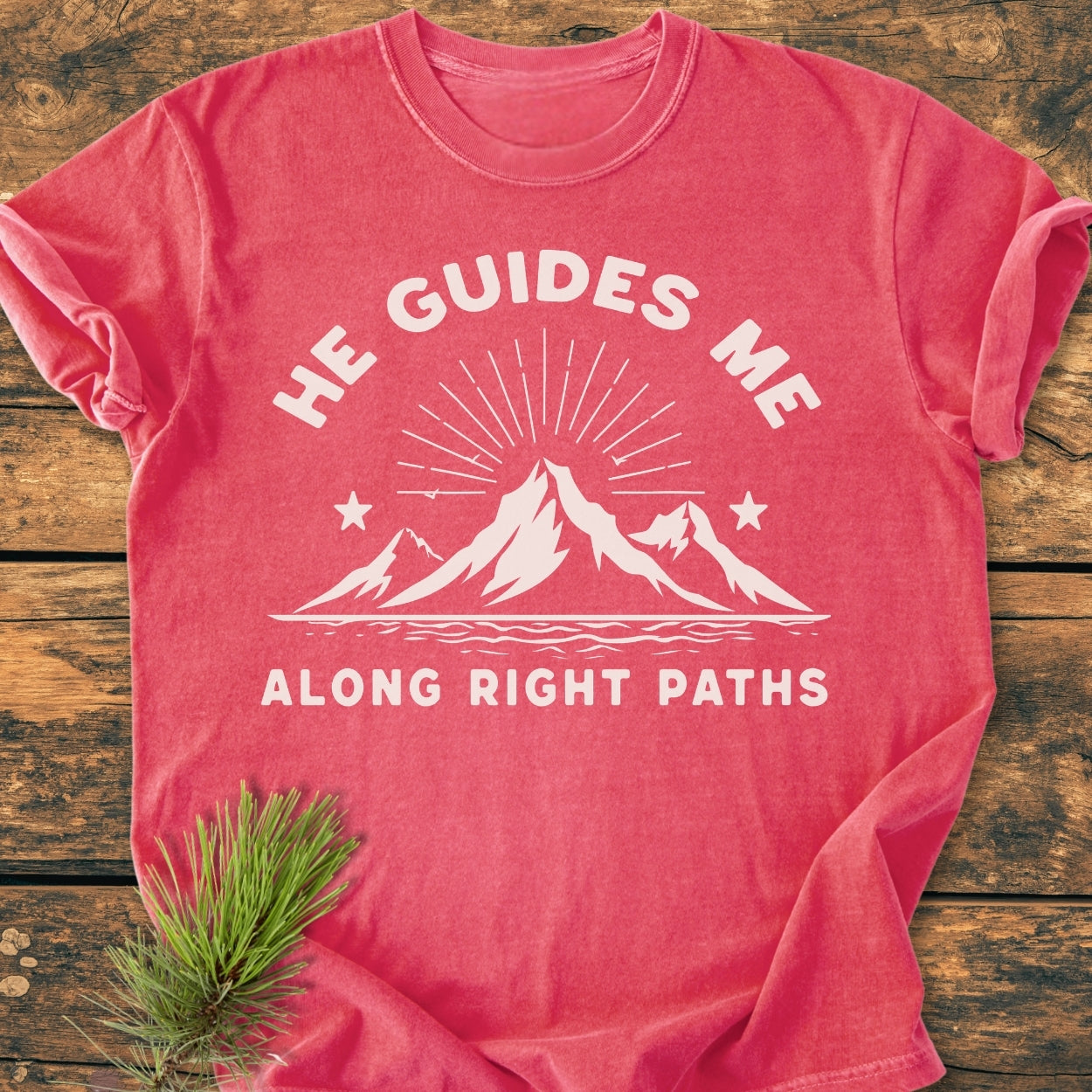 He Guides Me - Vintage Wash Tee features a stylized mountain graphic and text, laid flat on wood, highlighting its soft, relaxed fit. A pine sprig complements the rustic presentation.
