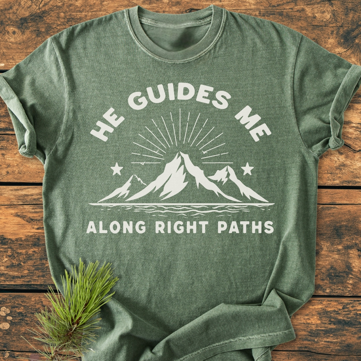 He Guides Me - Vintage Wash Tee displays a stylized mountain range with stars and text on a heather-green background, laid on wood, featuring soft, durable, garment-dyed cotton for daily comfort.