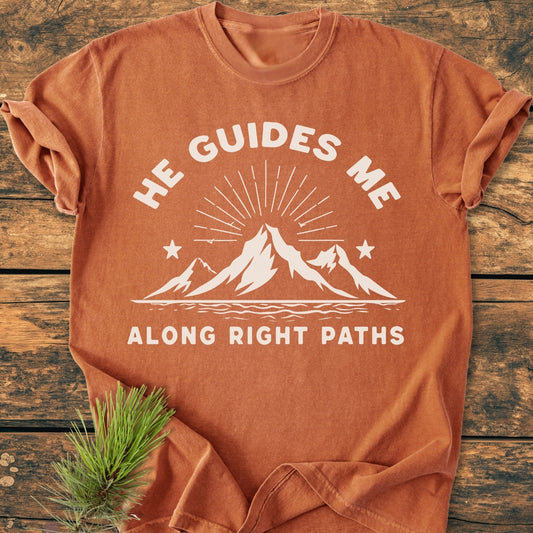 He Guides Me - Vintage Wash Tee features a white mountain graphic with sunbeams and stars on a rust-orange, short-sleeve T-shirt, laid on wood, highlighting its relaxed, durable design.