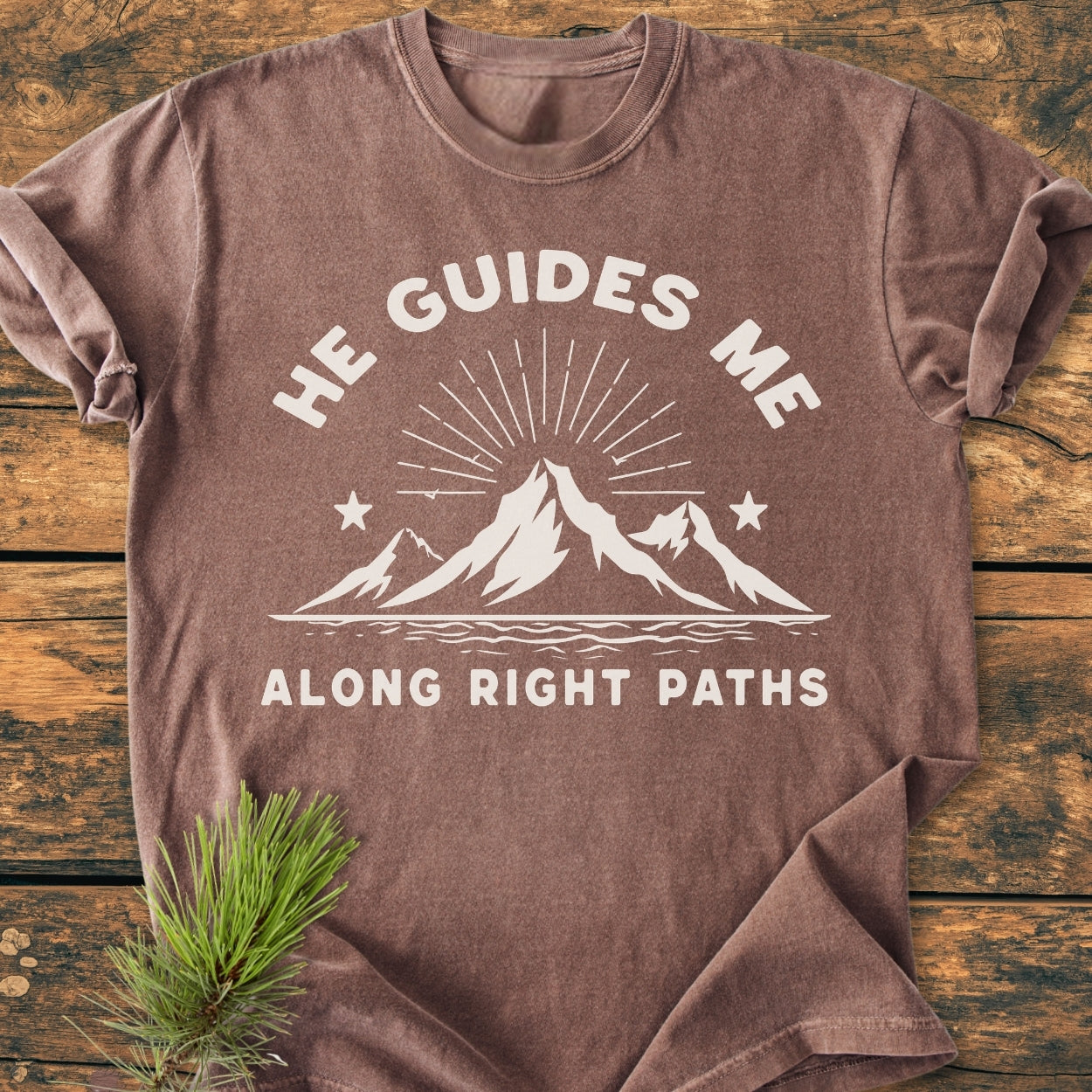 He Guides Me - Vintage Wash Tee: Earth-toned T-shirt featuring a white mountain graphic with sunbeams and stars, laid on wood with a pine sprig, emphasizing comfort and durability with a relaxed fit.