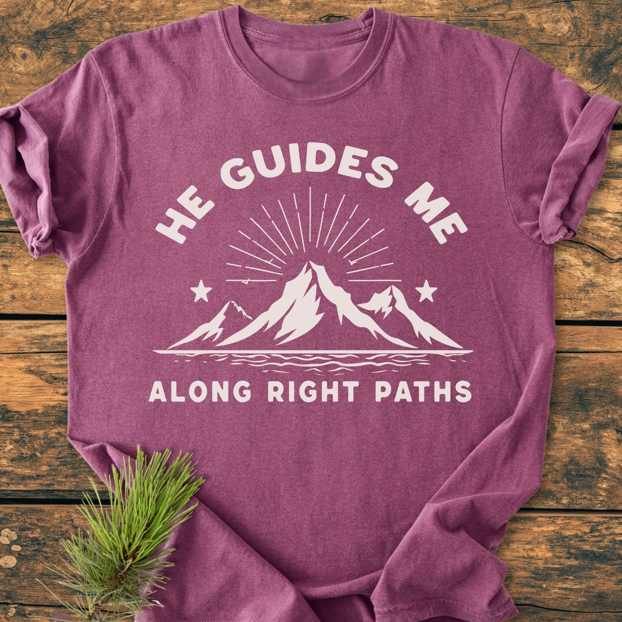 He Guides Me - Vintage Wash Tee features a white mountain graphic with text on a plum T-shirt, folded on rustic wood, showcasing its soft, relaxed fit and durable design, accented by a pine sprig.