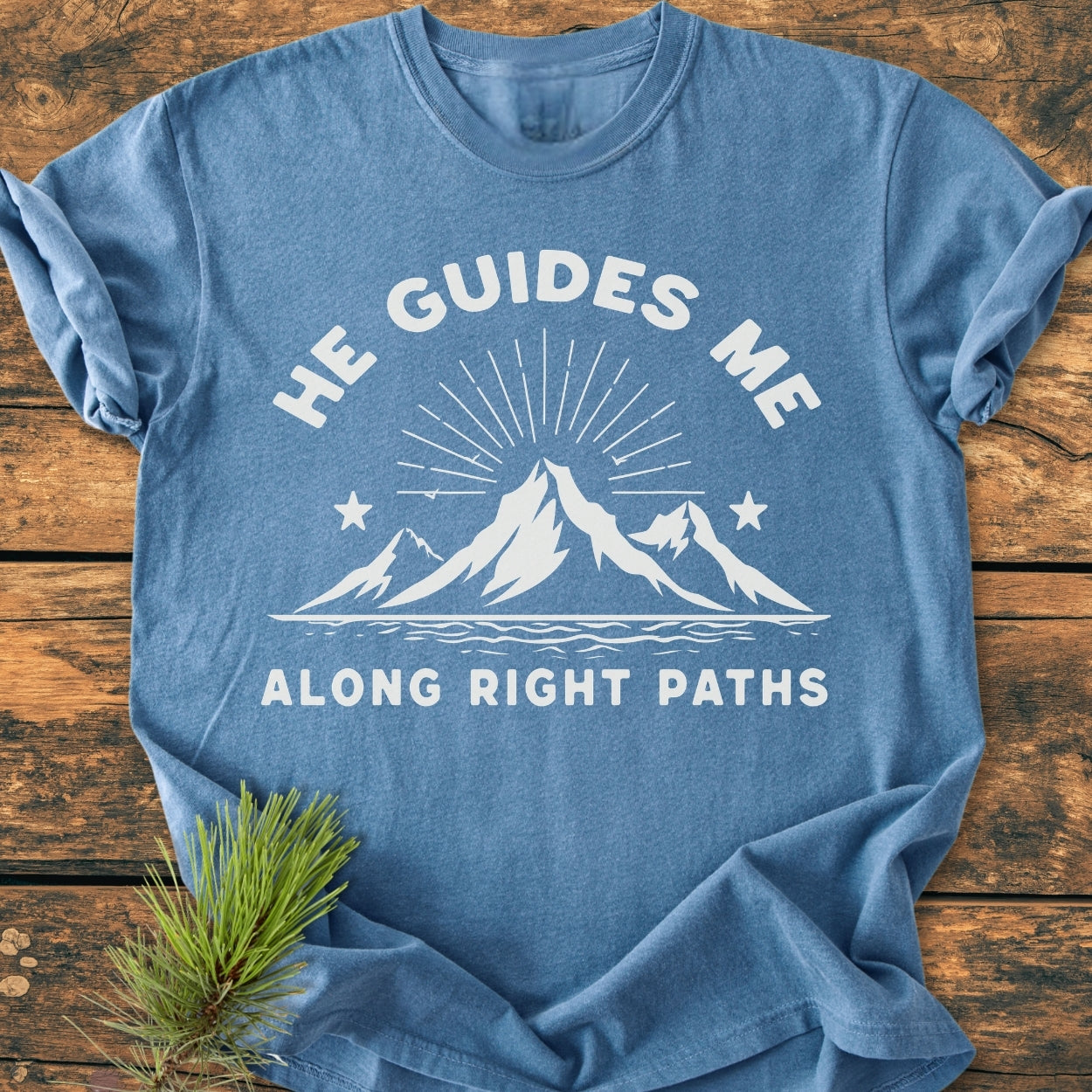 He Guides Me - Vintage Wash Tee features a stylized mountain range with sunbeams and water, screen-printed in white. Laid flat with a pine bough accentuating its rustic appeal.