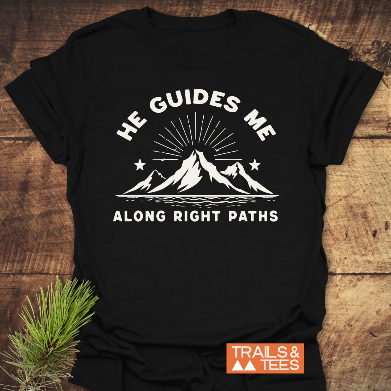 He Guides Me T-Shirt featuring a stylized mountain range and sun-rays graphic with motivational text. Laid on wood, it symbolizes outdoor adventure, crafted for comfort and durability by Trails & Tees.