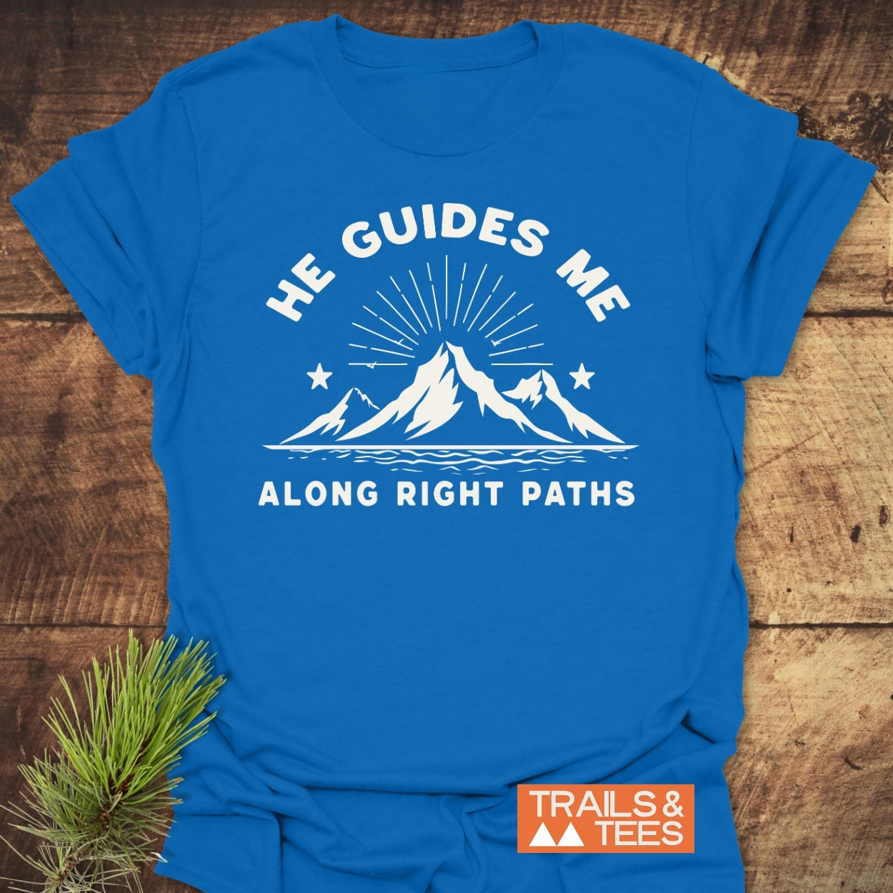 He Guides Me T-Shirt with a mountain and sunburst design, featuring HE GUIDES ME and ALONG RIGHT PATHS text, laid on wood with a Trails & Tees logo and pine sprig nearby.