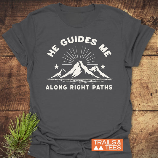 He Guides Me T-Shirt featuring a stylized mountain range with sun-rays and stars printed on the front, laid on wood. Includes a small orange TRAILS & TEES logo in the corner.