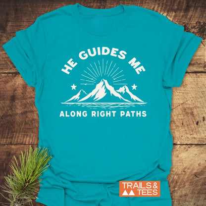He Guides Me T-Shirt features a white mountain graphic and inspirational text on a turquoise-blue backdrop, laid on wood with a pine sprig, emphasizing adventure-ready design.