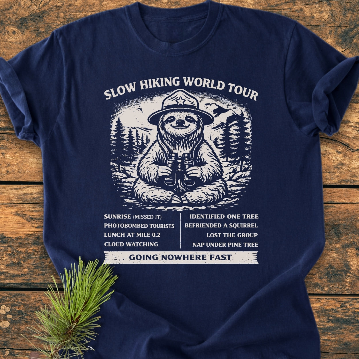 Slow Hiking World Tour - Vintage Wash Tee features a playful sloth in a ranger hat with binoculars, surrounded by pine trees and mountains, accompanied by a humorous faux itinerary and Going Nowhere Fast tagline.