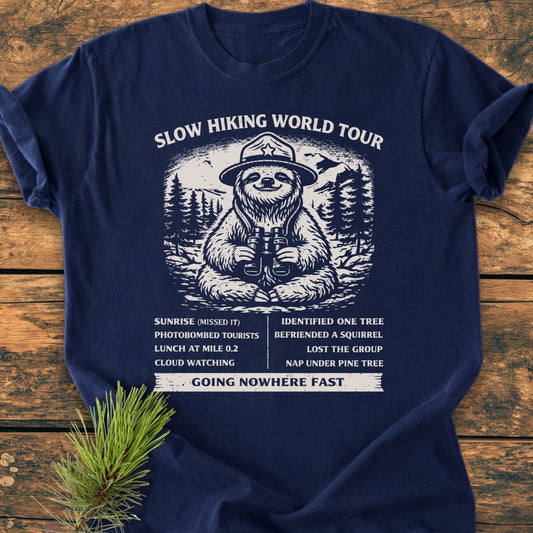 Slow Hiking World Tour - Vintage Wash Tee features a playful sloth in a ranger hat with binoculars, surrounded by pine trees and mountains, accompanied by a humorous faux itinerary and Going Nowhere Fast tagline.