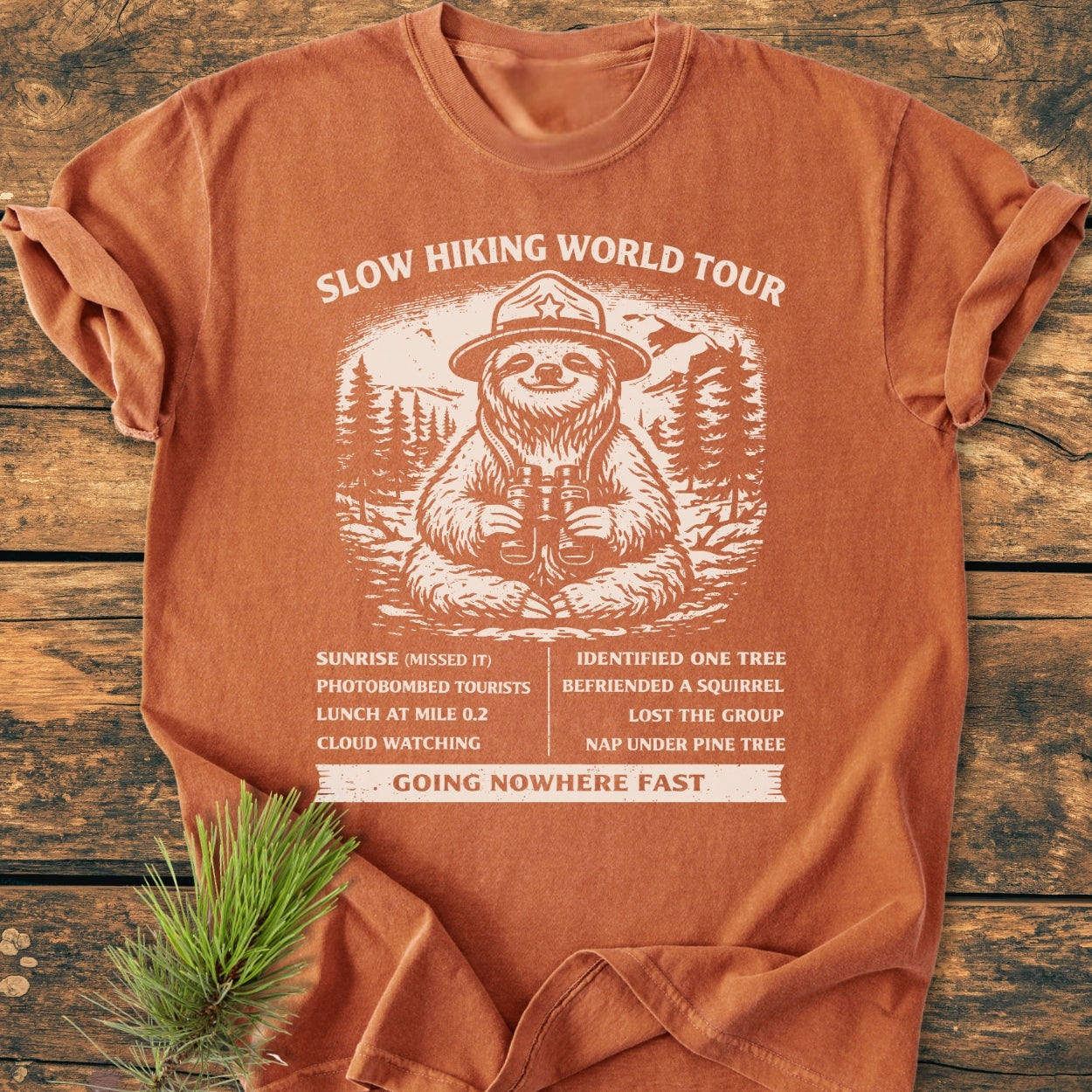 Slow Hiking World Tour - Vintage Wash Tee featuring a sloth graphic with a humorous itinerary, displayed flat on wood with pine sprig. Comfortable, garment-dyed, ring-spun cotton with relaxed fit.