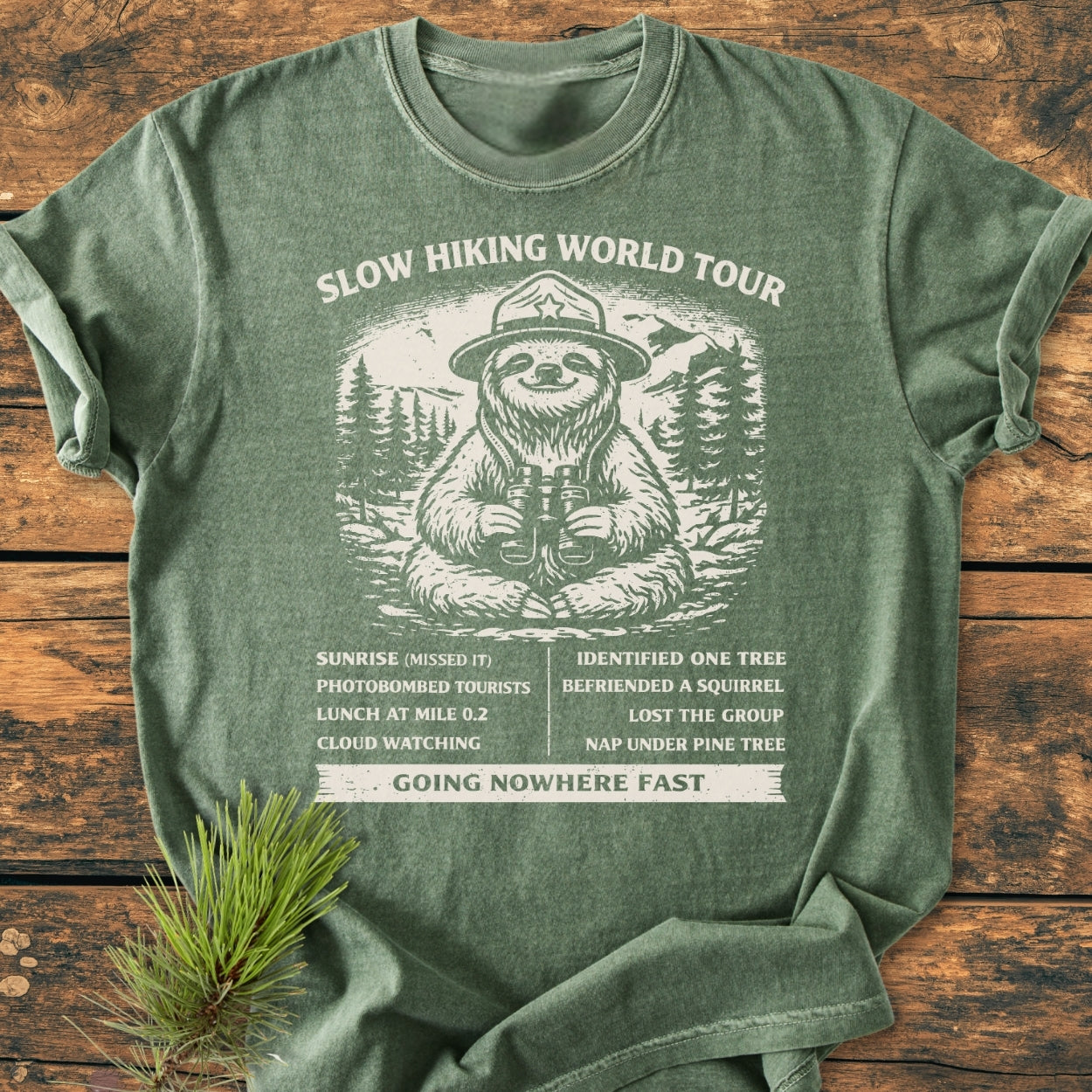 The Slow Hiking World Tour - Vintage Wash Tee features a playful sloth hiker design with a humorous itinerary, printed on a soft, green, short-sleeved T-shirt laid flat.