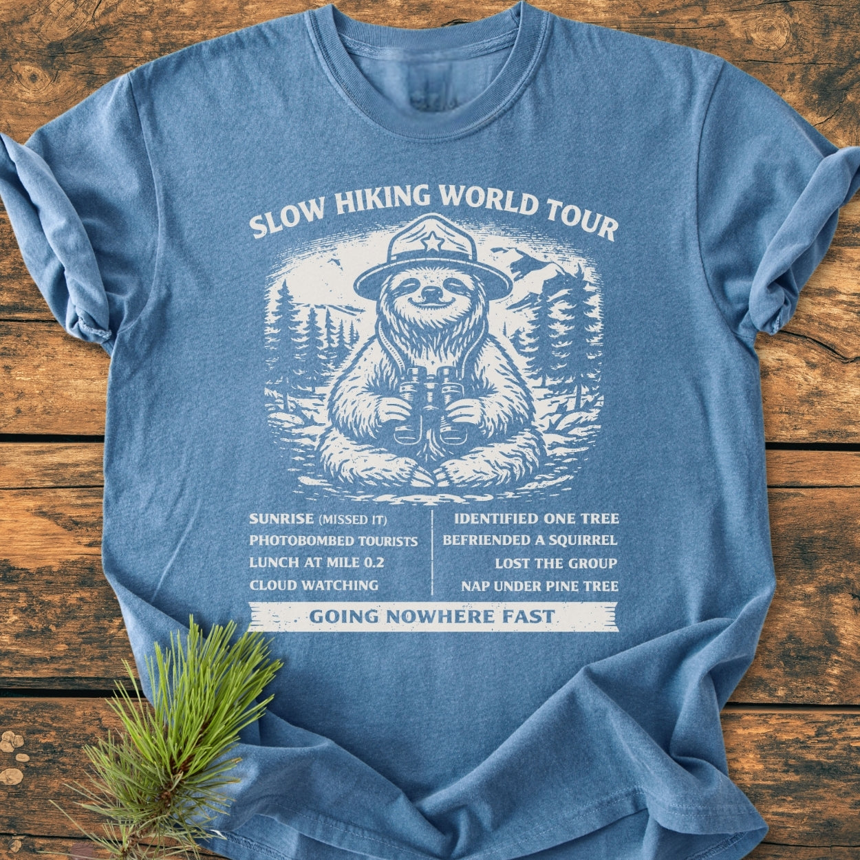 Slow Hiking World Tour - Vintage Wash Tee features a sloth in a ranger hat with binoculars, surrounded by trees and mountains, capturing a humorous itinerary and GOING NOWHERE FAST tagline on heather-blue fabric.