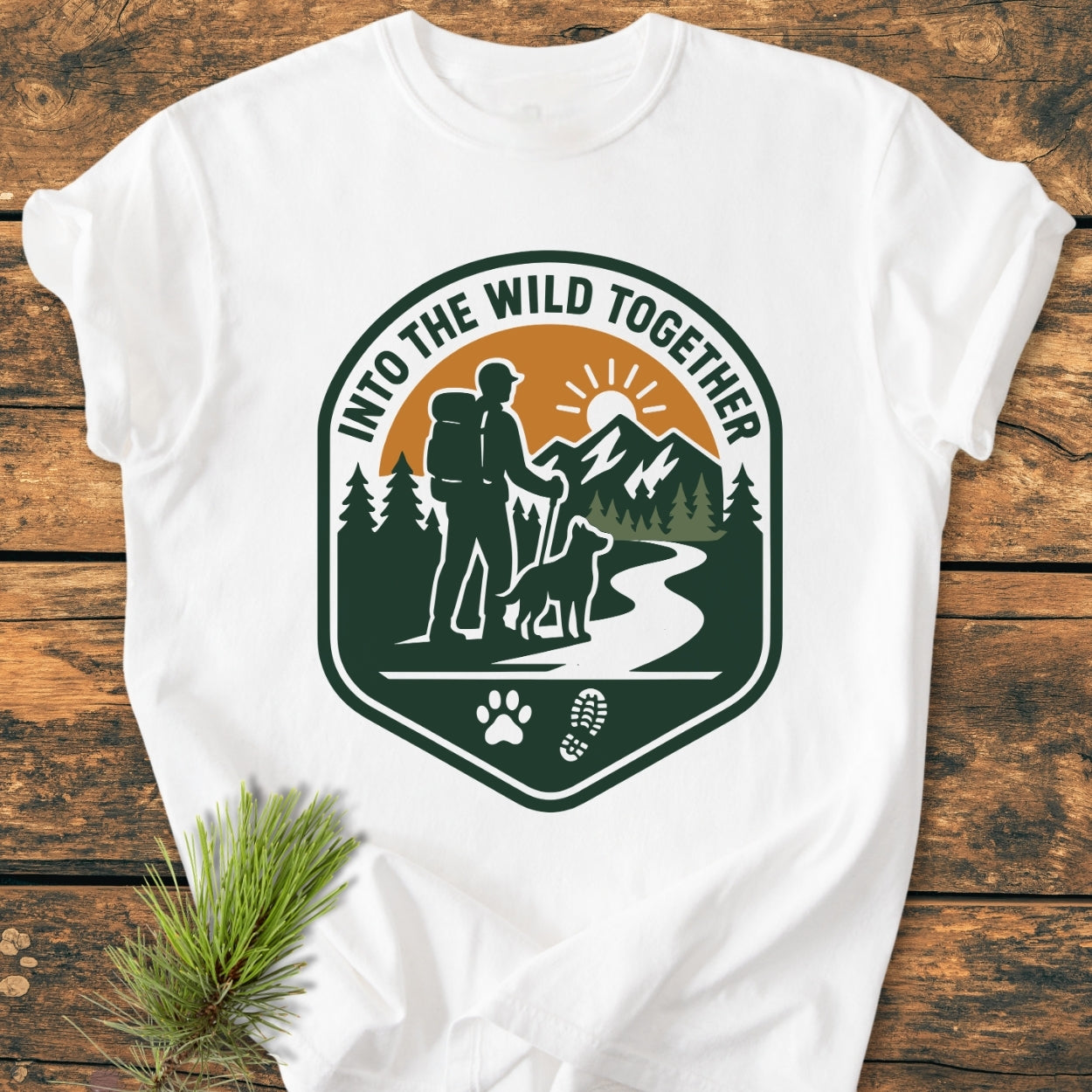 Into The Wild Together - Vintage Wash Tee featuring a badge graphic of a hiker and dog on a trail, printed on a soft, durable, garment-dyed white T-shirt laid on a wooden surface.