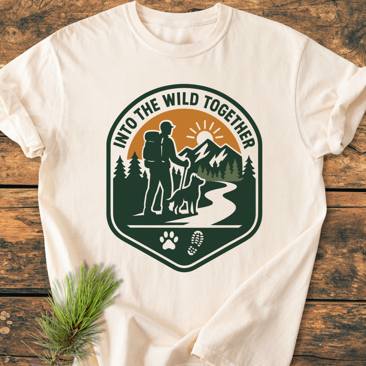 Into The Wild Together - Vintage Wash Tee features a badge graphic with a hiker, dog, trail, and sunlit mountains, celebrating adventure; laid flat on a wooden surface, showcasing its relaxed fit.