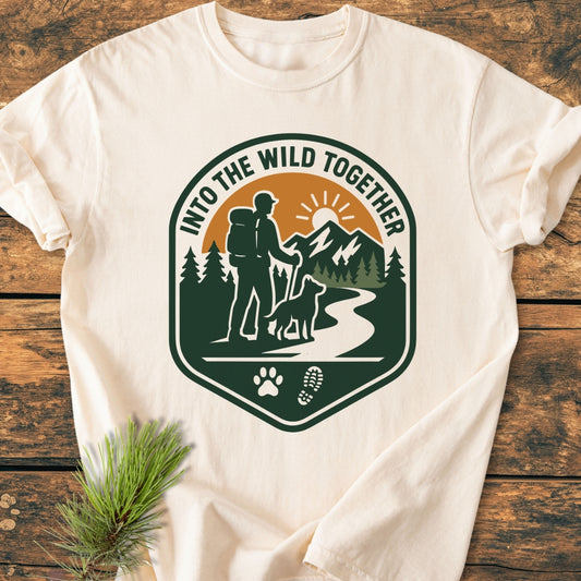 Into The Wild Together - Vintage Wash Tee features a badge graphic with a hiker, dog, trail, and sunlit mountains, celebrating adventure; laid flat on a wooden surface, showcasing its relaxed fit.
