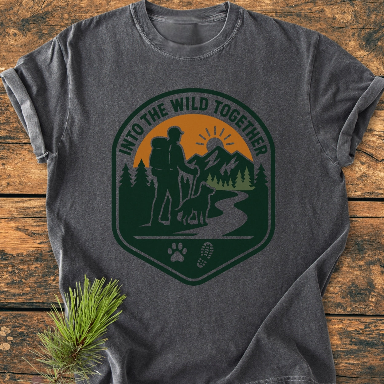 Into The Wild Together - Vintage Wash Tee featuring a badge design with a hiker and dog on a trail, printed on a charcoal-gray Comfort Colors shirt, styled with a pine sprig.