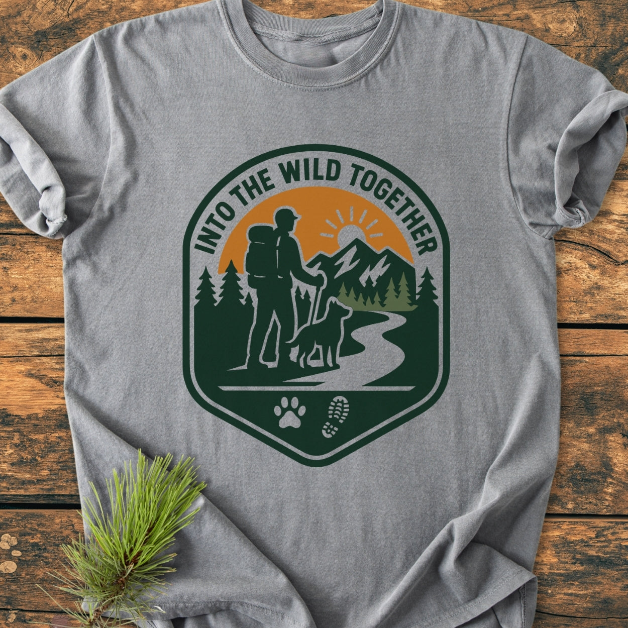 Into The Wild Together - Vintage Wash Tee featuring a backpacker and dog graphic, with a scenic trail, pine trees, mountain, and sun, laid flat on rustic wood with a pine sprig accent.