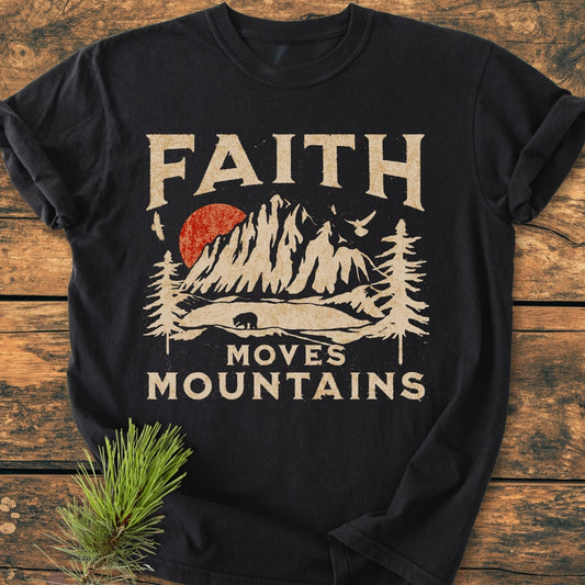 Faith Moves Mountains - Vintage Wash Tee: Black t-shirt with a stylized mountain graphic, red sun, pine trees, bear silhouette, and birds, featuring bold “FAITH MOVES MOUNTAINS” lettering, flat-laid on wood.