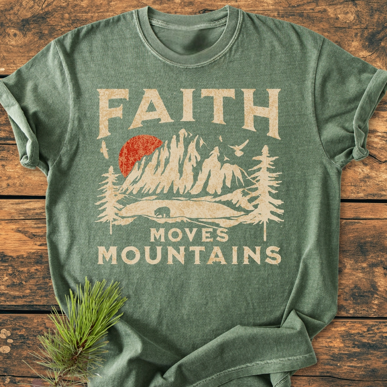 Faith Moves Mountains - Vintage Wash Tee featuring a distressed mountain graphic with a bear and pine trees, laid flat on rustic wood. Crafted from cozy, soft-washed, garment-dyed, 100% ring-spun cotton.