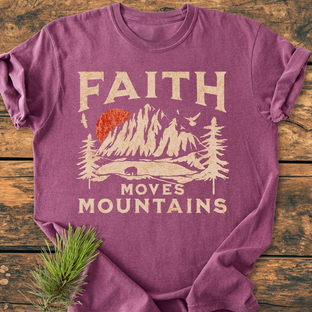 Faith Moves Mountains - Vintage Wash Tee features a heather-purple T-shirt with FAITH and a mountain scene, including a bear silhouette and MOVES MOUNTAINS text, styled on a rustic wooden surface.