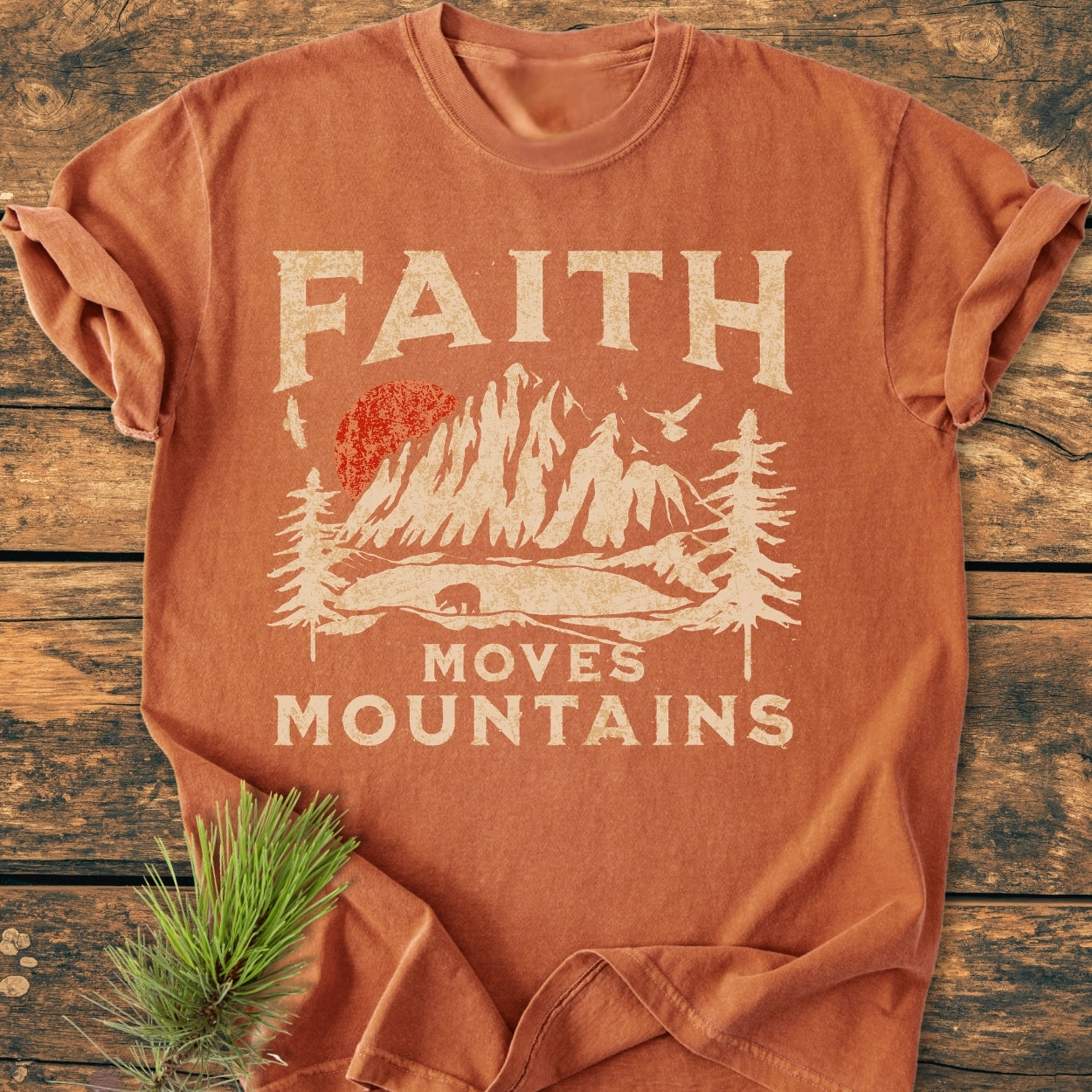 Faith Moves Mountains - Vintage Wash Tee features a rust-orange hue, stylized mountain scene with a bear and slogan, laid flat on wood with a pine sprig accentuating its rustic charm.