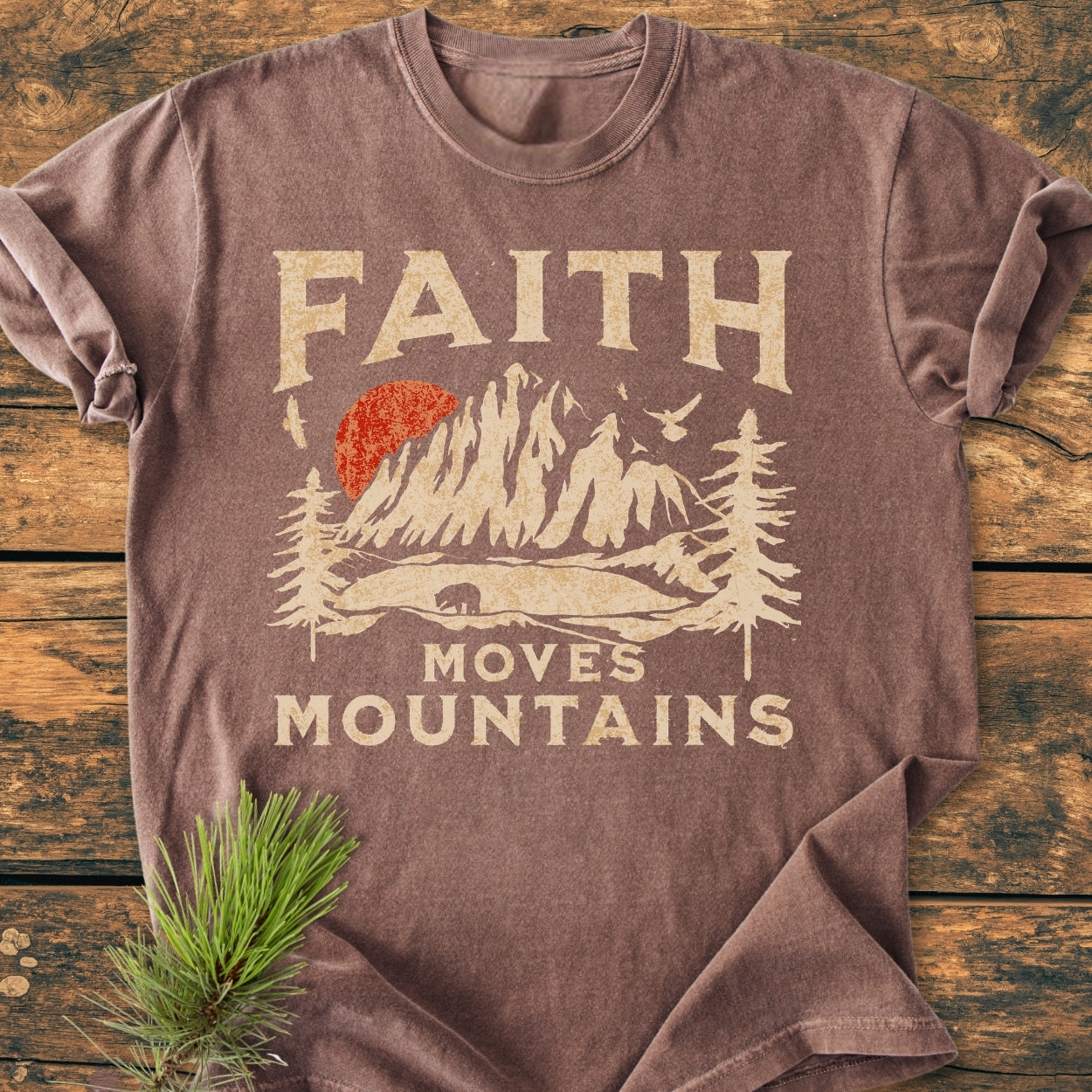 Faith Moves Mountains - Vintage Wash Tee: Brown, vintage-washed t-shirt with a stylized mountain and sun graphic, FAITH MOVES MOUNTAINS text, laid on rustic wood with decorative pine branch.