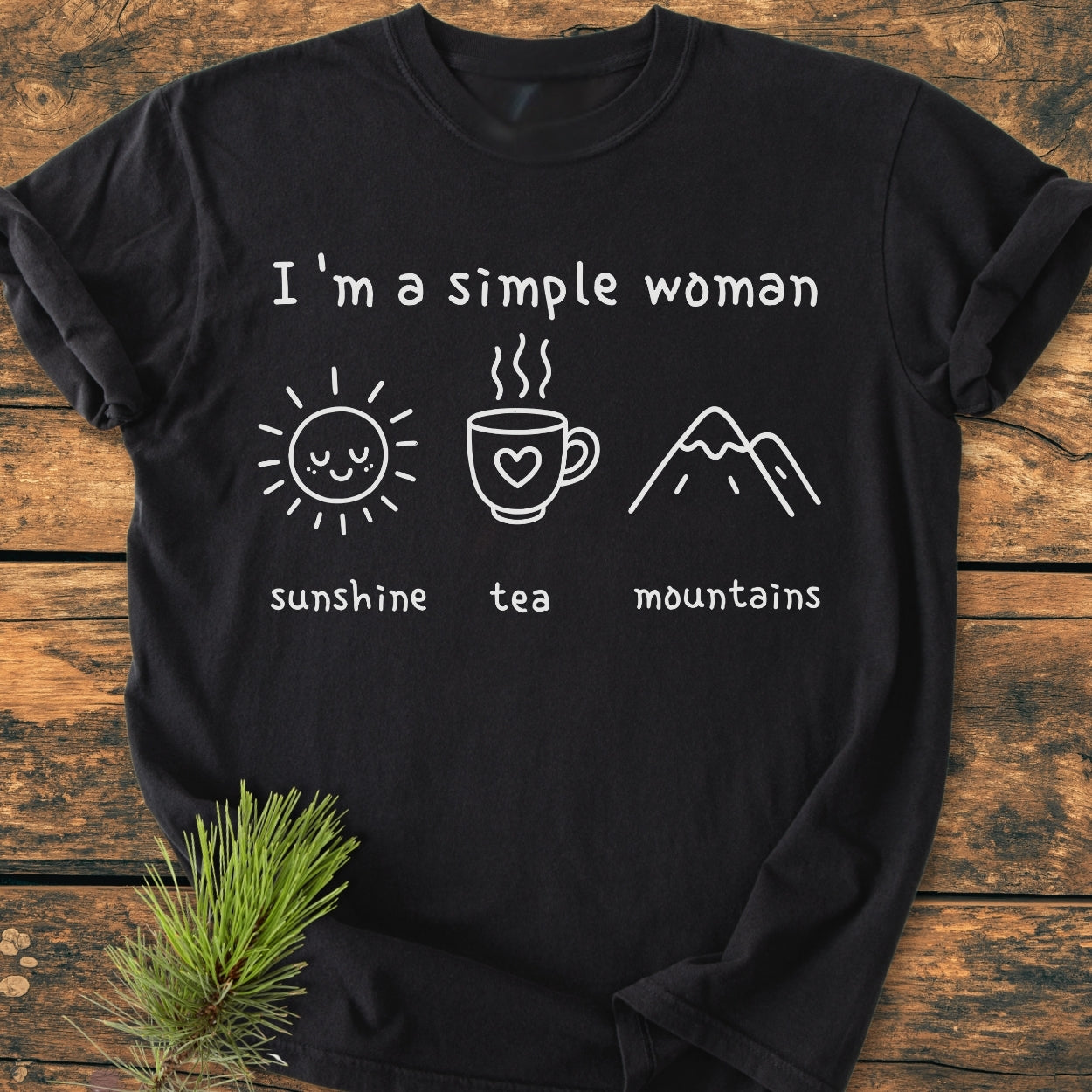I'm A Simple Woman. Sunshine, Tea, Mountains - Vintage Wash Tee featuring white icons of a sun, mug, and mountains on black. Laid on wood, showcasing its relaxed fit and durable construction.