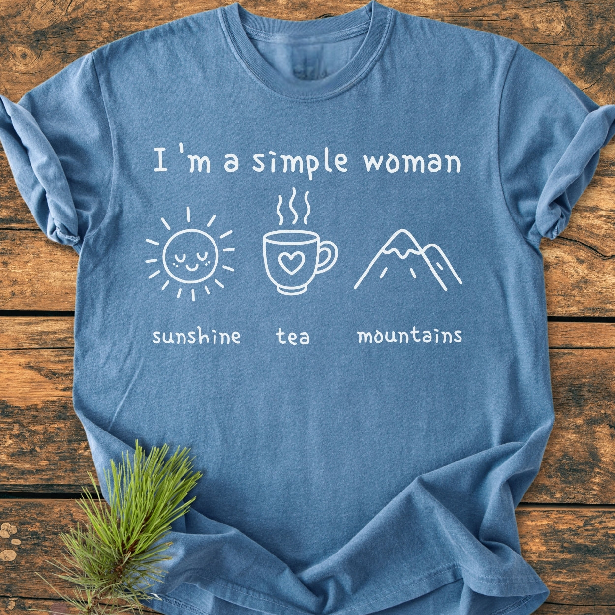 I'm A Simple Woman. Sunshine, Tea, Mountains - Vintage Wash Tee, featuring hand-drawn icons of a sun, mug, and mountains, laid flat on a wooden surface.