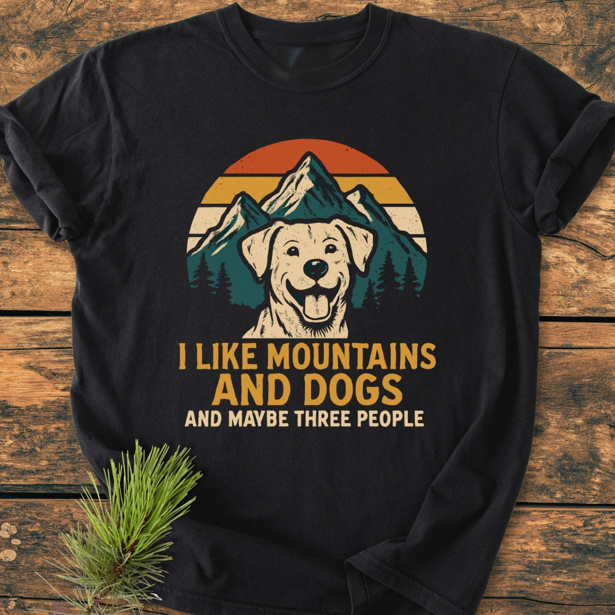 Black I Like Mountains And Dogs - Vintage Wash Tee with retro graphic of a dog's face, mountains, and sunset. Features slogan: I LIKE MOUNTAINS AND DOGS AND MAYBE THREE PEOPLE. Laid on wooden surface.