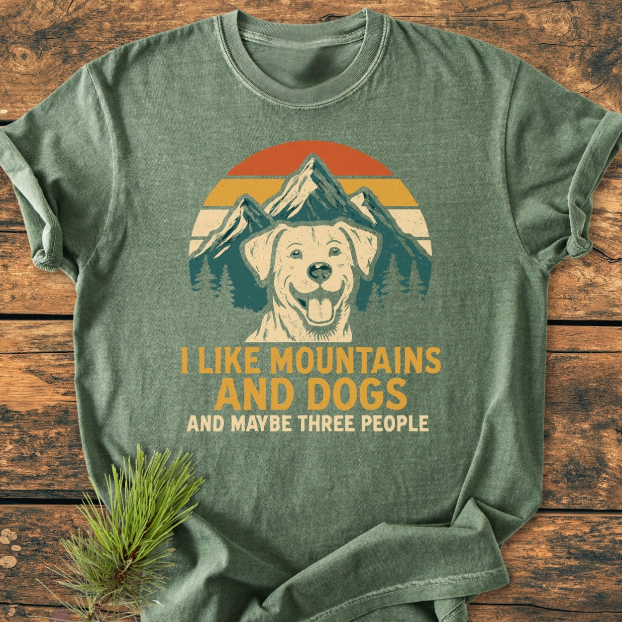 I Like Mountains And Dogs - Vintage Wash Tee displayed on a wooden surface, featuring a dog graphic with mountains and sunset stripes, complemented by the slogan, highlighting its relaxed fit and durable stitching.