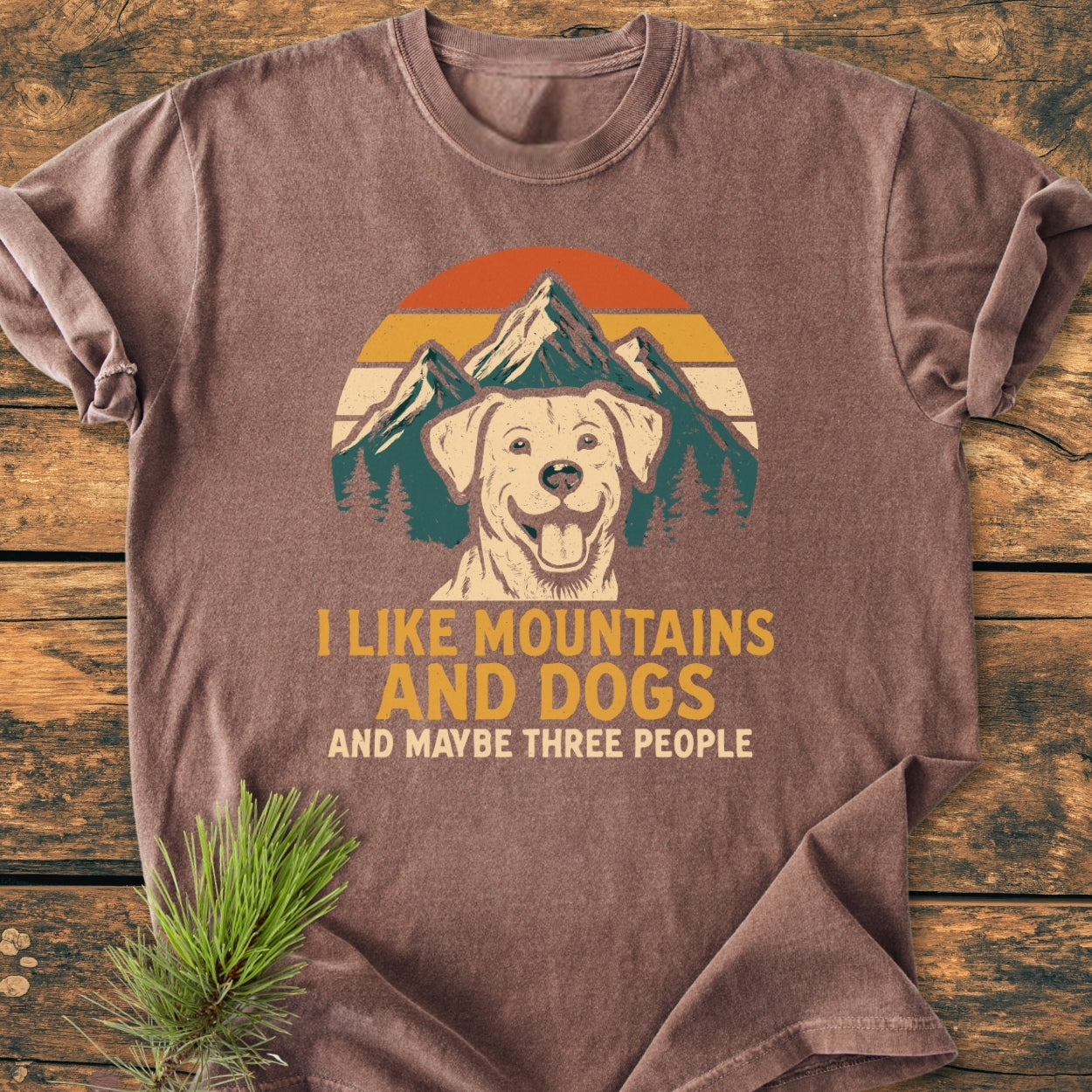 I Like Mountains And Dogs - Vintage Wash Tee: Brown T-shirt with a vintage graphic of snow-capped mountains and a smiling dog, featuring playful text, laid flat on a wooden background.
