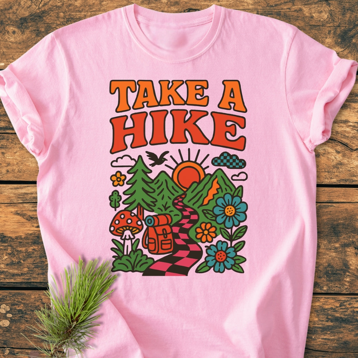 Take A Hike Funky - Vintage Wash Tee on rustic wood, featuring a retro outdoor graphic with trail, mountains, backpack, trees, wildflowers, mushroom, bird, and clouds. Cozy, relaxed fit, garment-dyed cotton.